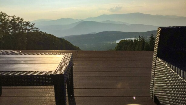 A wooden deck with a table and a wicker chair, overlooking a mountainous landscape. - Pension Wildflowers (Tateshina)