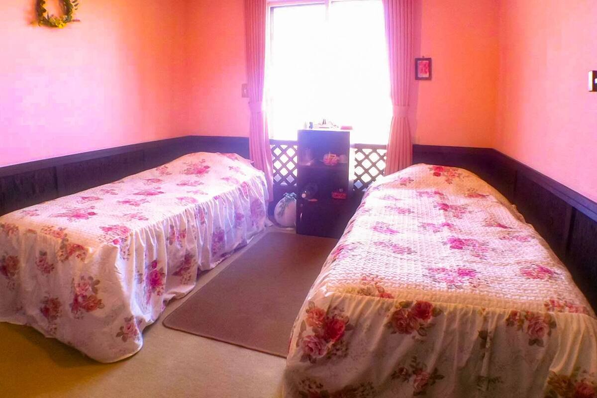 Ein Zimmer mit zwei Einzelbetten, jeweils mit einem blumigen Überwurf, einem kleinen Tisch und einem fenster mit Vorhängen.