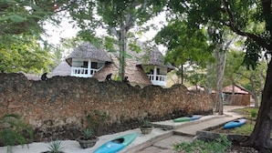 Un camino lleva a cabañas con techo de paja y una pared de piedra, rodeado de macetas con plantas y un kayak azul.