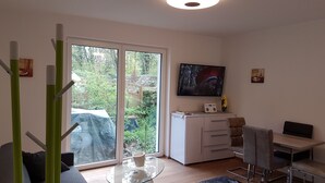 A modern living room with a large sliding glass door leading to a garden, a flat-screen TV mounted on the wall, a small white cabinet, and a dining area with a table and chairs. - Luxusapartment Steinbreche (Bergisch Gladbach)
