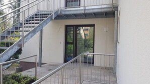 A building with a metal staircase leading to a door, a balcony with a railing, and a view of trees and buildings in the background. - Luxusapartment Steinbreche (Bergisch Gladbach)