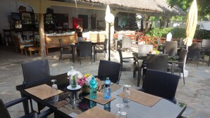 An outdoor dining area with wicker furniture and a thatched roof. Tables are set with placemats, water bottles, and glasses. - Morning Star Diani (Diani Beach)