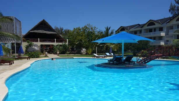 A clear blue swimming pool with lounge chairs and umbrellas, surrounded by a resort-style building with a thatched roof and palm trees. - Morning Star Diani (Diani Beach)