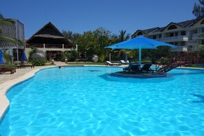 Una piscina azul cristalina con sillas de descanso y sombrillas, rodeada por un edificio de estilo resort con techo de paja y palmeras.