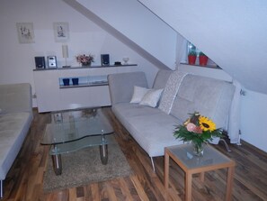 A modern living room with a glass coffee table, a grey sofa, a wooden side table with a vase of flowers, and a kitchen area with white cabinets and a red pot on the shelf. - Großzügiges Zweiraumapartment Troisdorf (Troisdorf)