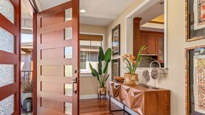 A hallway with a wooden door, a console table with decorative items, and a view into a kitchen area. - Stunning 4BR KaMilo Home 349 at Mauna Lani – Relaxed Elegance & Pool/Spa (Kamuela)