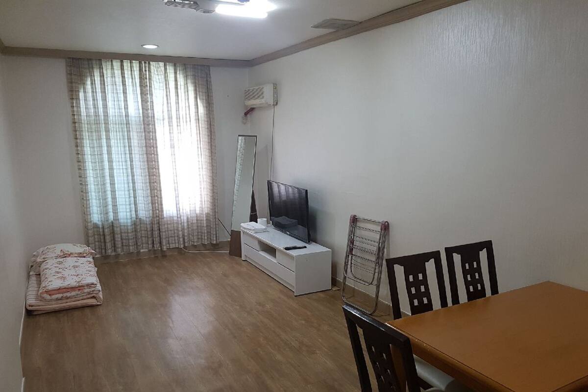 A living room with a wooden floor, a TV on a white stand, a dining table with chairs, and a window with curtains.