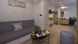 Ein modernes Wohnzimmer mit einem grauen Sofa, einem kleinen Tisch mit Frühstücksutensilien, einem an der Wand befestigten Fernseher und einer Küche im Hintergrund.