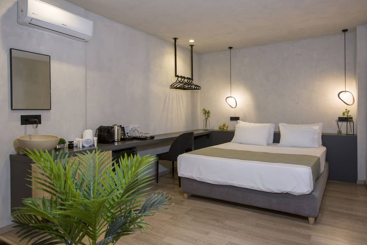 A modern hotel room with a large bed, a desk, a chair, a small table with a plant, and an air conditioning unit.