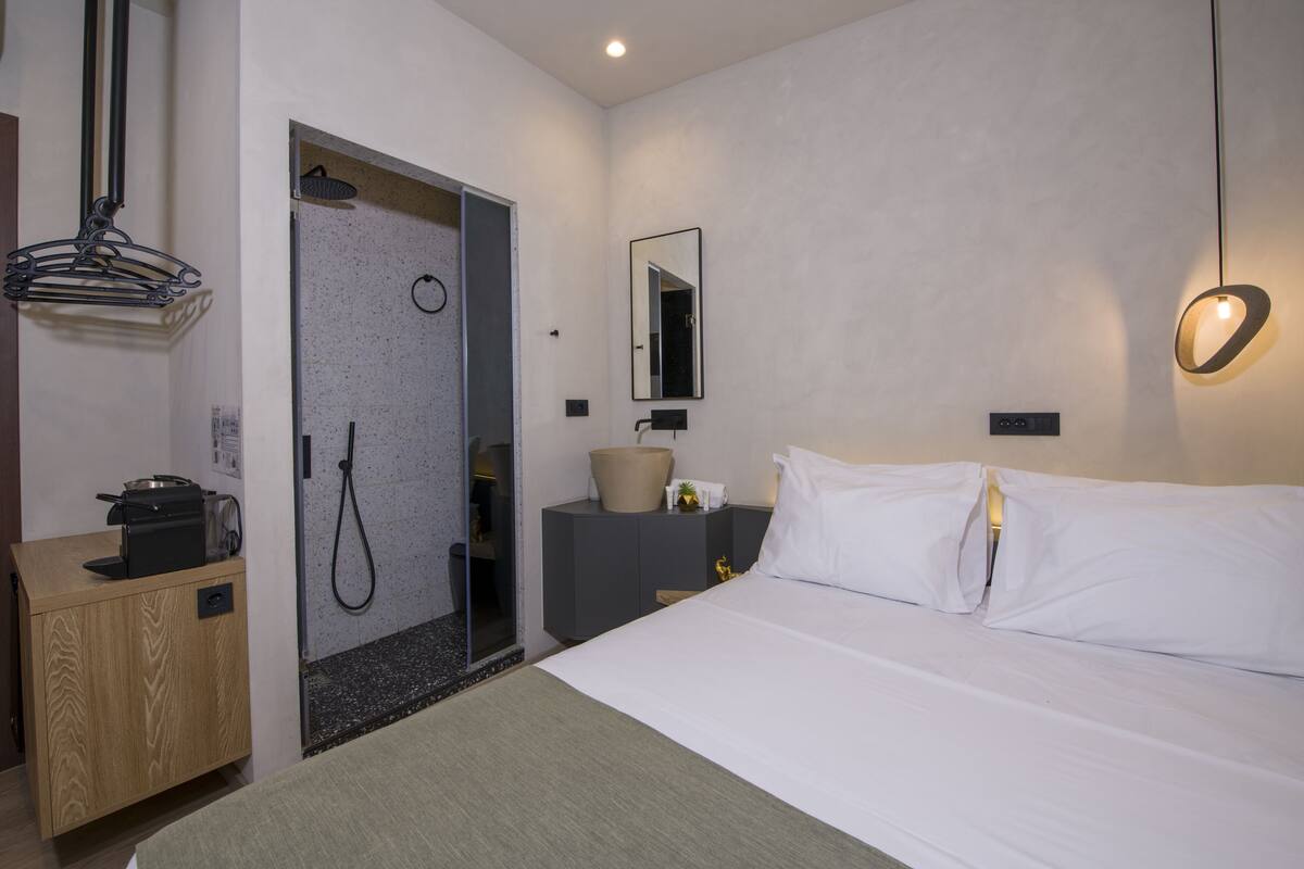 A hotel room with a bed, a wooden side table, a bathroom with a shower, a mirror, and a wall-mounted light fixture.