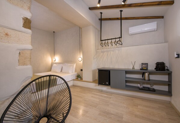 Une chambre moderne avec un lit, une table de chevet et une chaise. On y trouve une étagère intégrée avec une machine à café et une bouilloire.