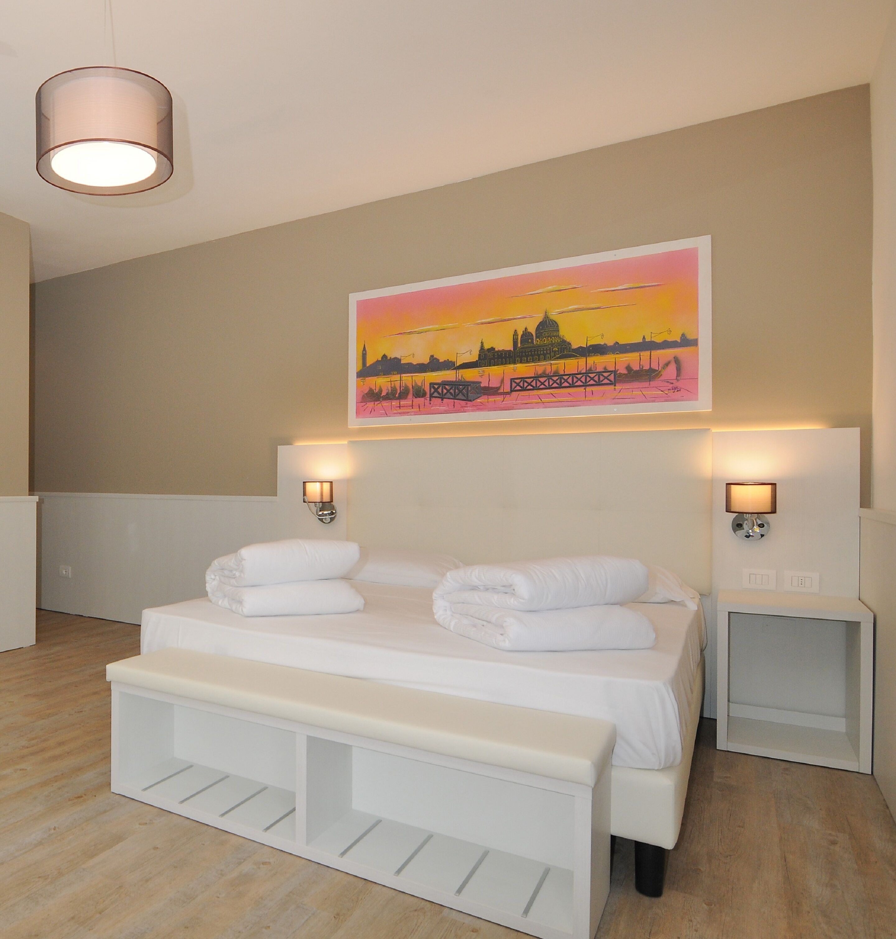 Habitación de hotel con cama, mesitas de noche, un banco y un cuadro con un paisaje urbano al atardecer.