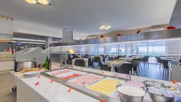 Zona de bufé con diversas opciones de comida, espacio de cocina con tostadora y zona de estar con mesas y sillas.