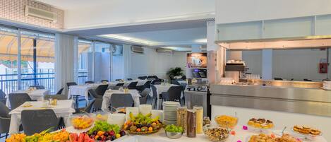 A buffet setup with various fruits, pastries, and beverages on a table in a spacious dining area with large windows and a kitchen in the background.