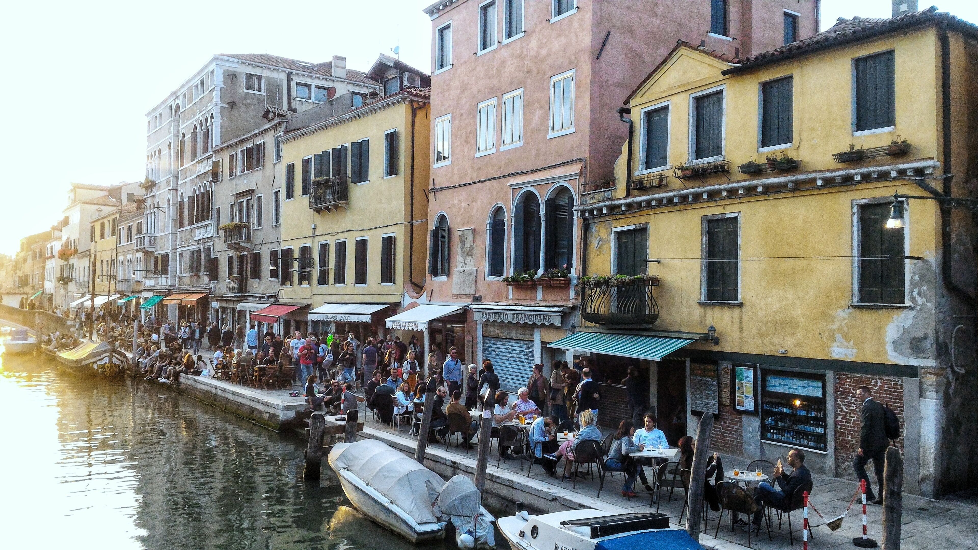 A canal-side street with buildings, outdoor seating, and boats.