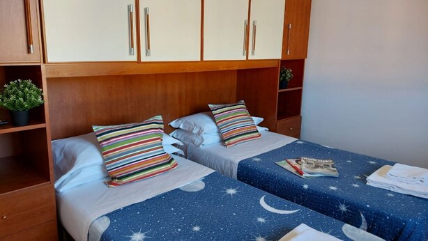 Ein Hotelzimmer mit zwei Betten, jeweils mit blauer Bettdecke mit Sternenhimmel-Muster. Darüber befinden sich Holzschränke und auf der linken Seite ein kleiner Topf mit Pflanze.