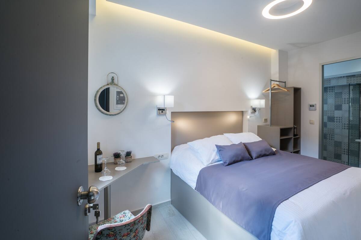 A modern hotel room with a bed, bedside table, mirror, and a small seating area.
