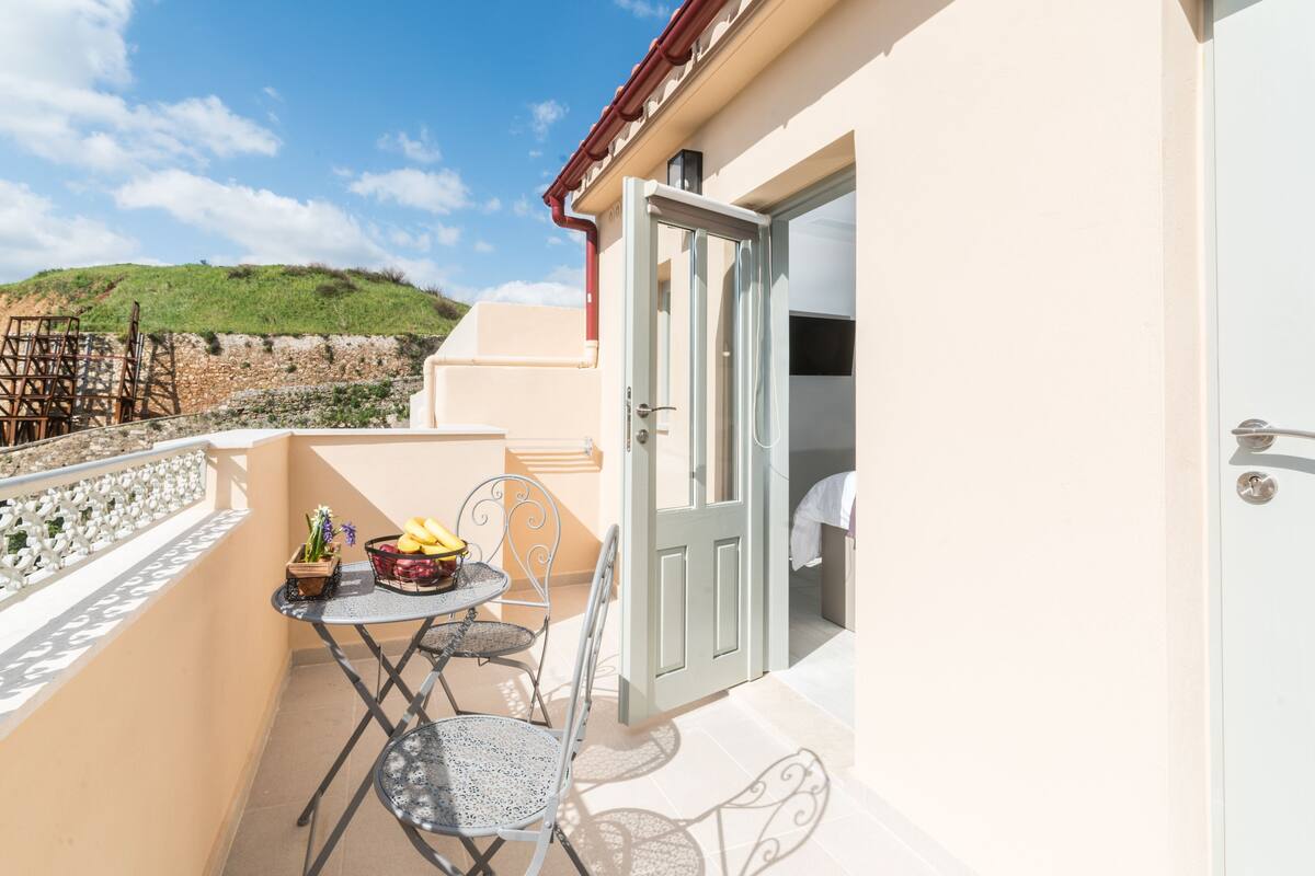 A balcony with a table and chairs, a door, and a view of a hill.