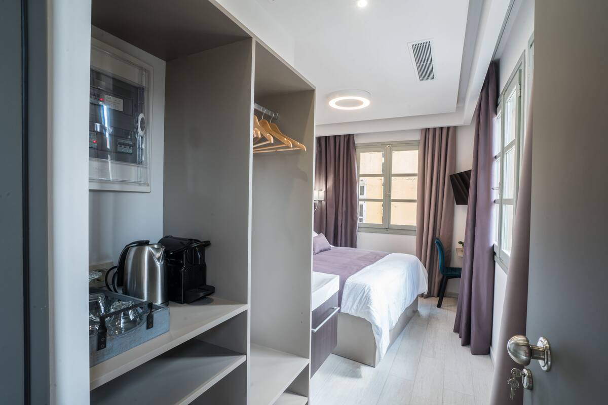 A hotel room with a bed, a wardrobe, a desk with a kettle, and a window with curtains.
