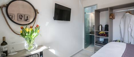 Una habitación de hotel moderna con una cama, un televisor montado en la pared, una mesita con un florero y un armario con ropa colgada.
