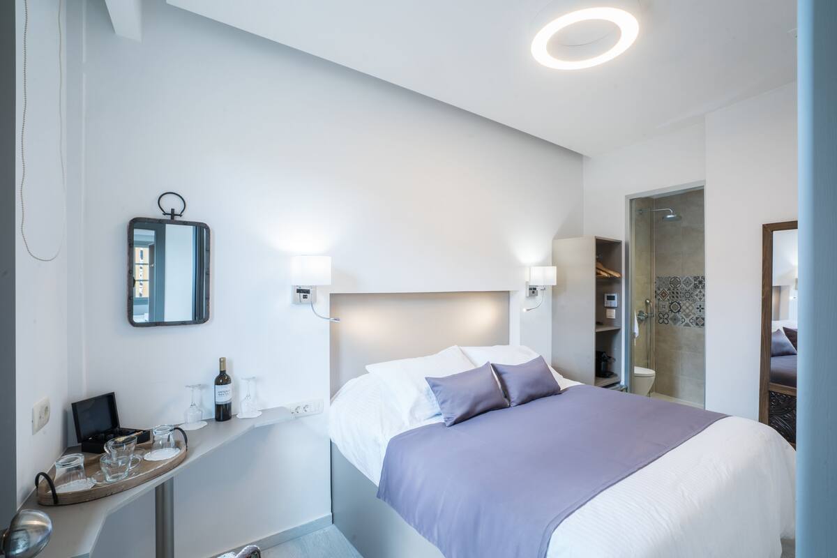 A modern hotel room with a neatly made bed, a bedside table with a wine bottle and glasses, a mirror, and a shower area.