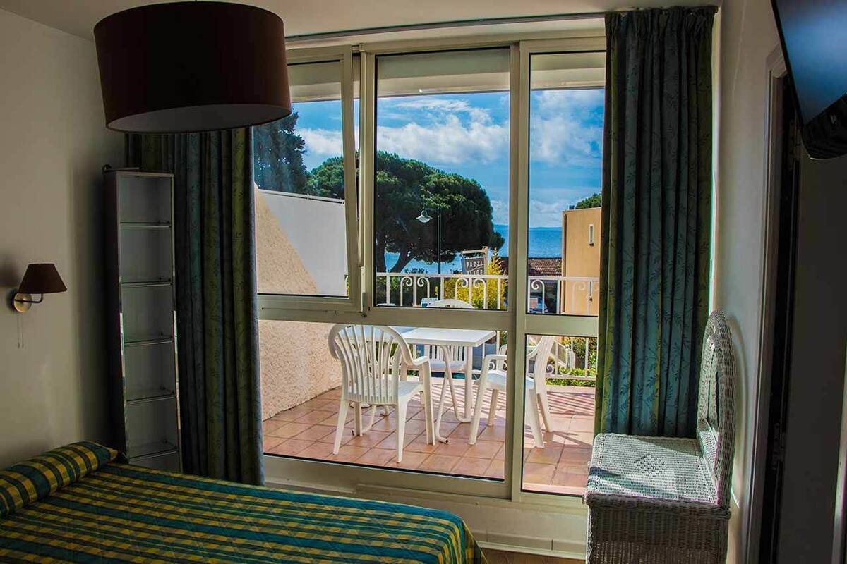A room with a bed, a window with a view of the sea, a chair, and a table outside.