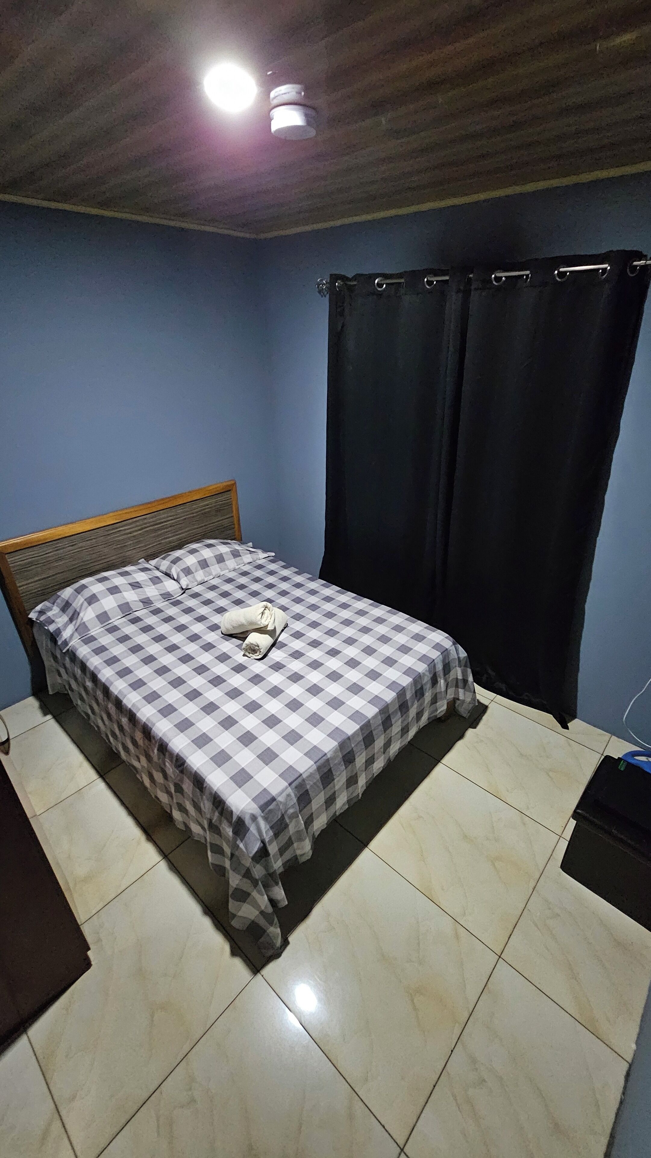 A bedroom with a checkered bedspread, a wooden headboard, a black curtain, and a tiled floor.