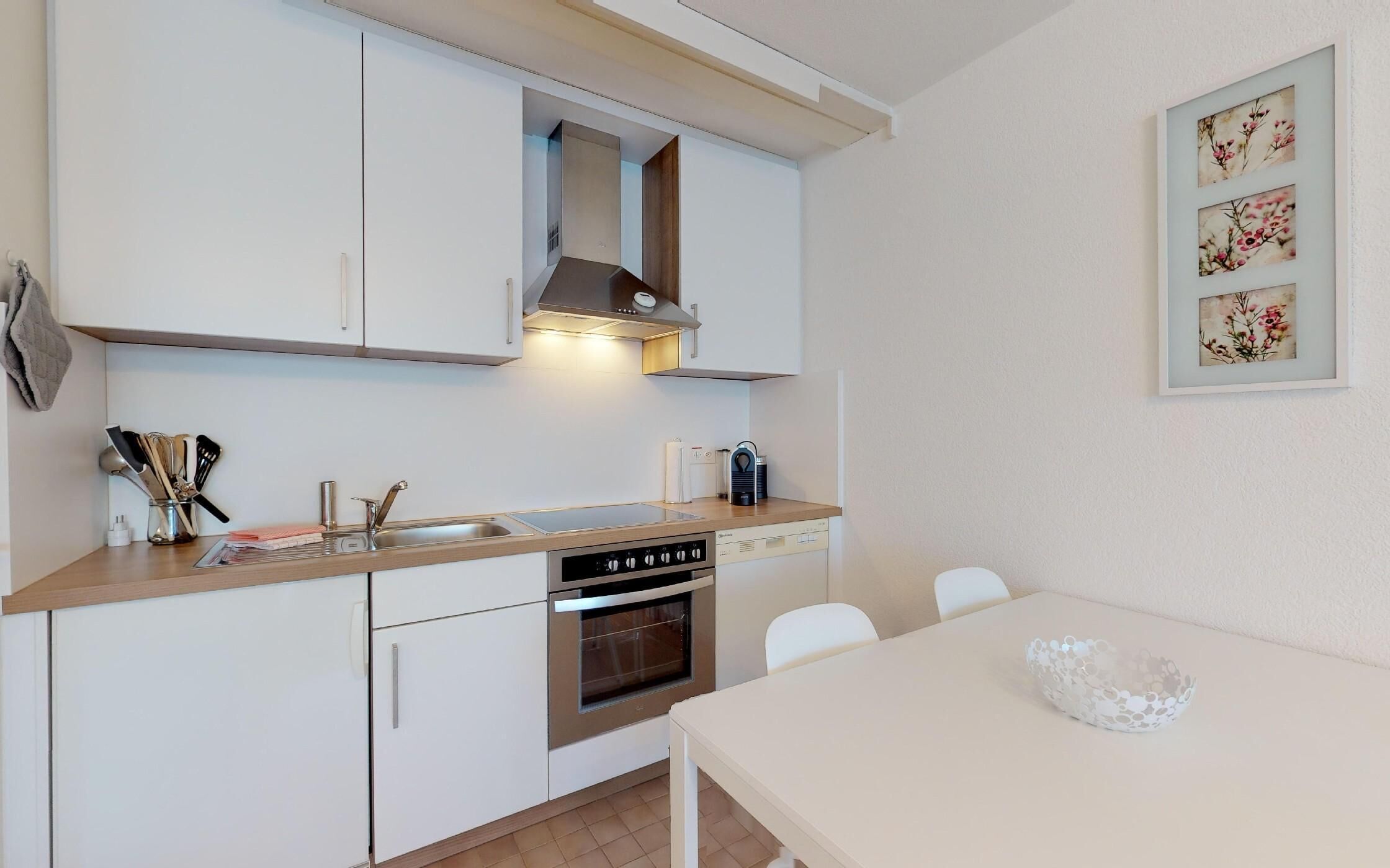 A compact kitchen with white cabinets, a built-in oven, a sink, and a dining area with a table and chairs.