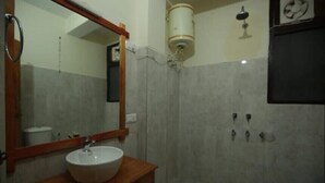 A bathroom with a wooden vanity, a white sink, a mirror, a shower with a wall-mounted light, and a toilet. - Jibhi Cottage (Banjar)