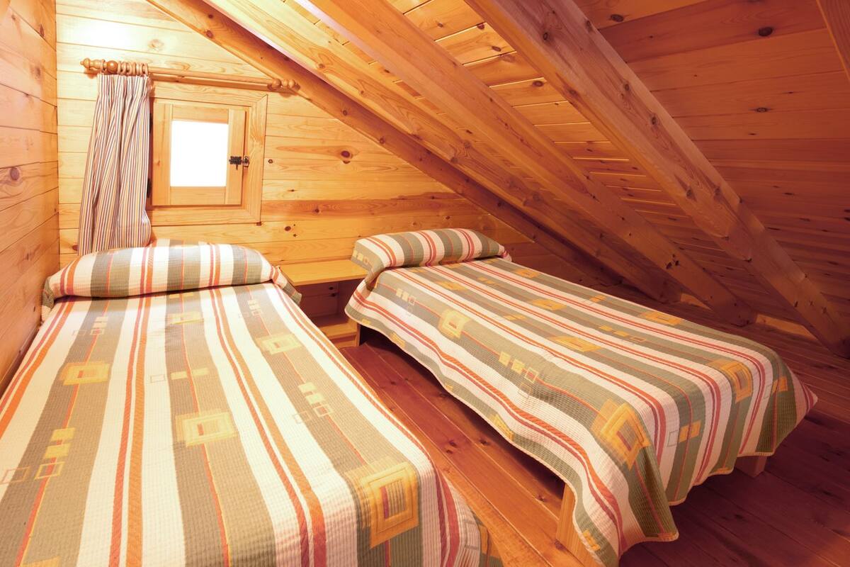 A cozy attic bedroom with two single beds, wooden walls, a sloped ceiling, and a small window.