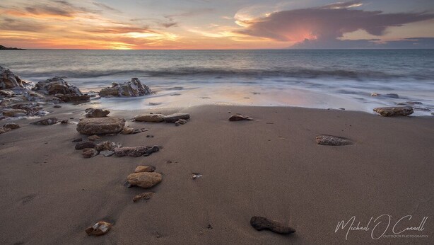 Ein Strand mit vereinzelten Felsen und einem Sonnenuntergang im Hintergrund.