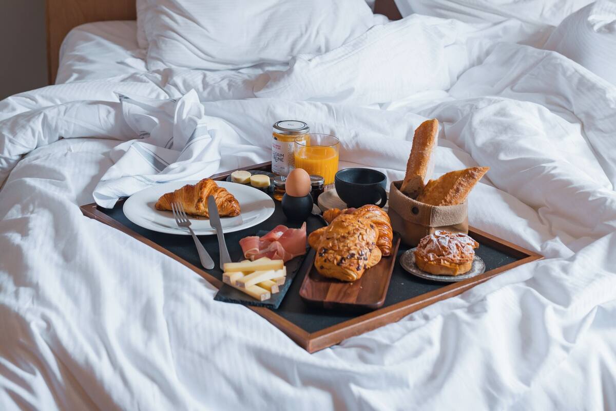 Una cama bien hecha con ropa de cama blanca, una bandeja con alimentos para el desayuno, incluyendo croissants, un croissant en un plato y un vaso de jugo de naranja.