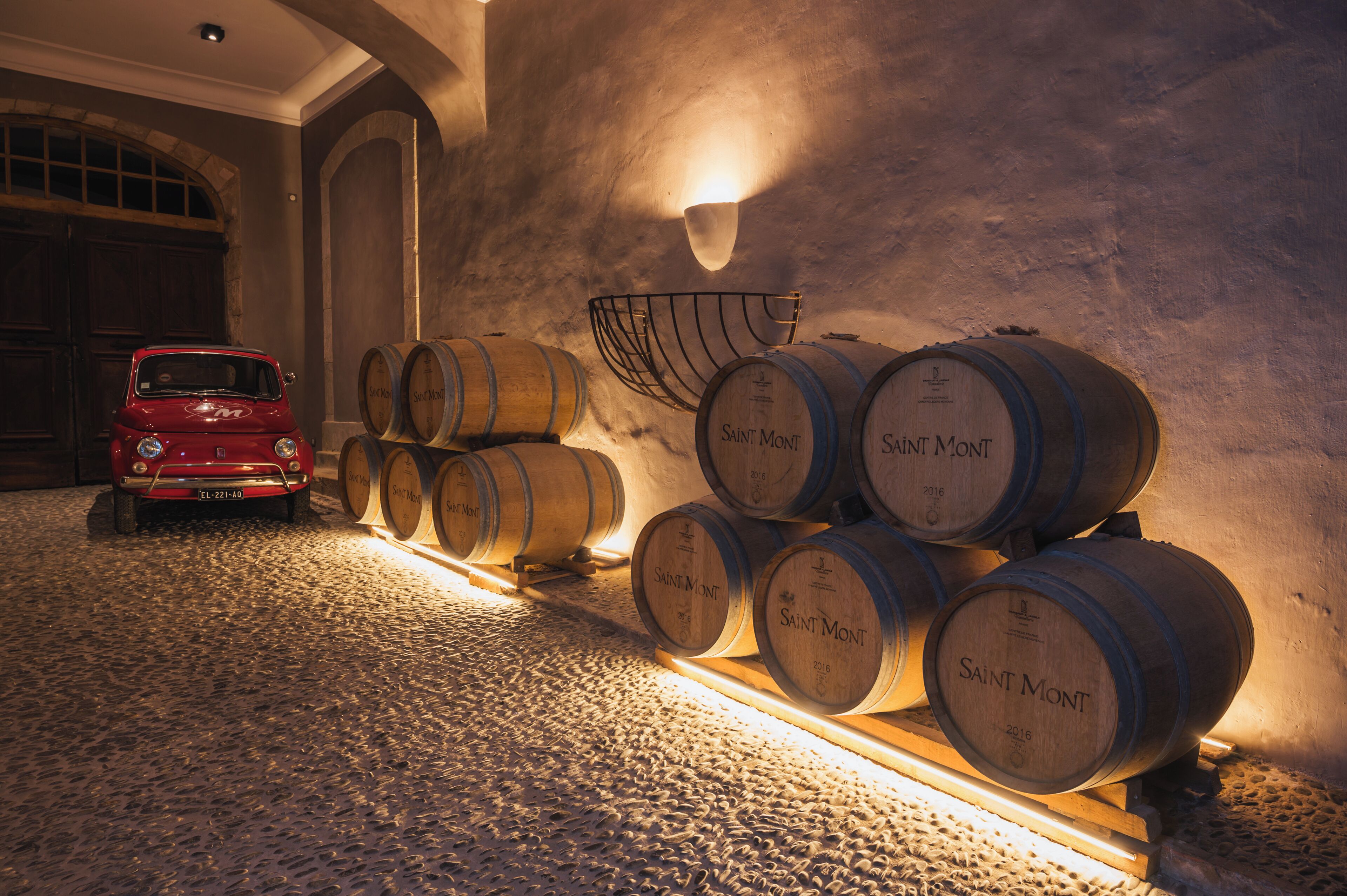 Une voiture ancienne et des tonneaux en bois remplis de vin se trouvent dans une pièce à la lumière tamisée, aux murs de pierre et au plafond en pente.
