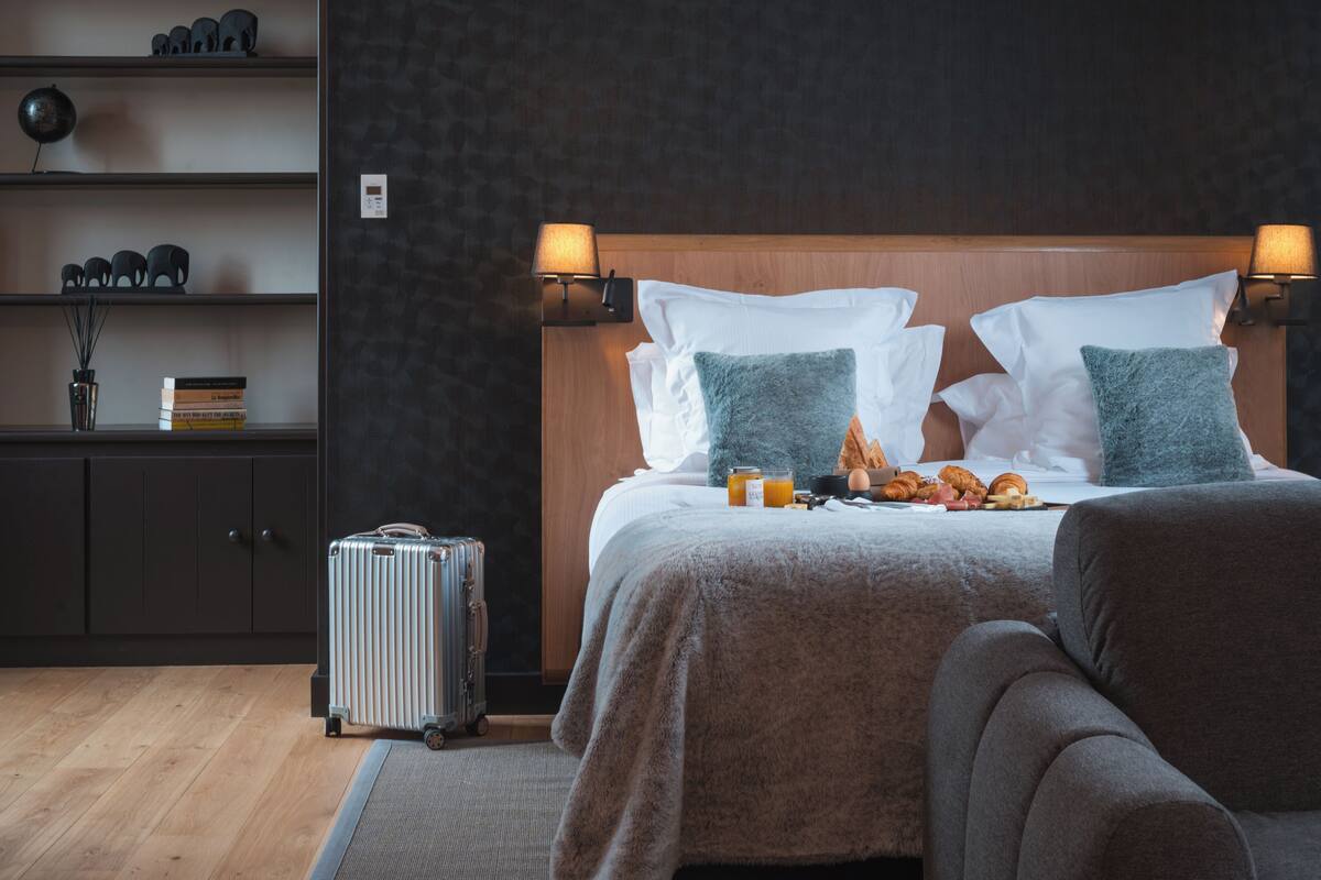 Habitación de hotel con cabecera de madera, dos lámparas de noche, cama con manta gris, dos almohadas, una maleta, estante con objetos decorativos y un reloj de pared.