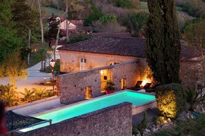 Un bâtiment en pierre doté d’une piscine, entouré d’arbres et avec une colline en arrière-plan.