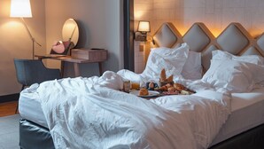 Ein ordentlich bezogenes Bett mit weißen Leinen, ein Nachttisch mit Lampe, ein Stuhl und ein Frühstücksbrett mit Croissants und Orangensaft.