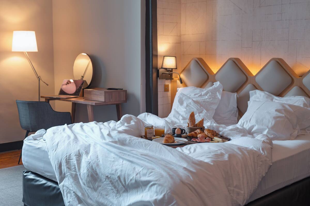 Una cama bien hecha con sábanas blancas, una mesita de noche con lámpara, una silla y una bandeja de desayuno con croissants y jugo de naranja.