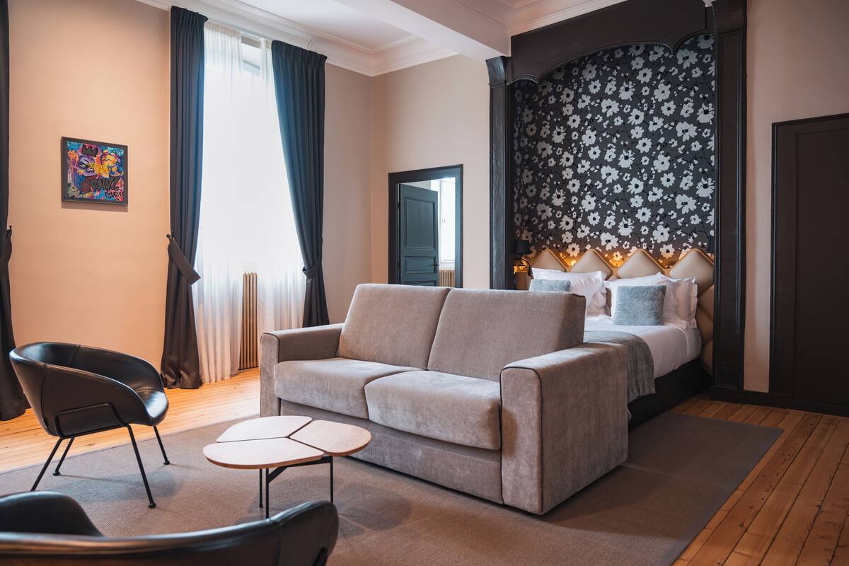Una habitación de hotel moderna con sofá, una mesita, una silla y una cama con cabecera floral.