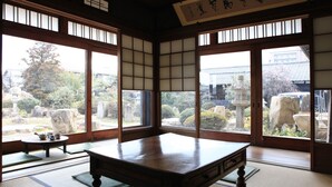 Una habitación tradicional japonesa con suelo de tatami, una mesa baja y grandes ventanales que dan a un jardín con rocas y un árbol.