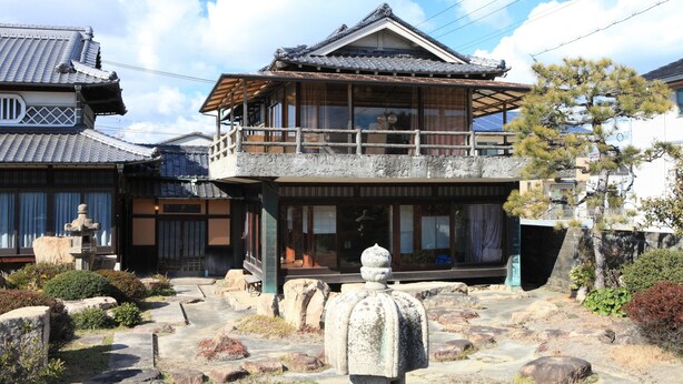 Un edificio japonés tradicional con balcón de madera, faroles de piedra y un jardín cuidado con rocas y árboles.