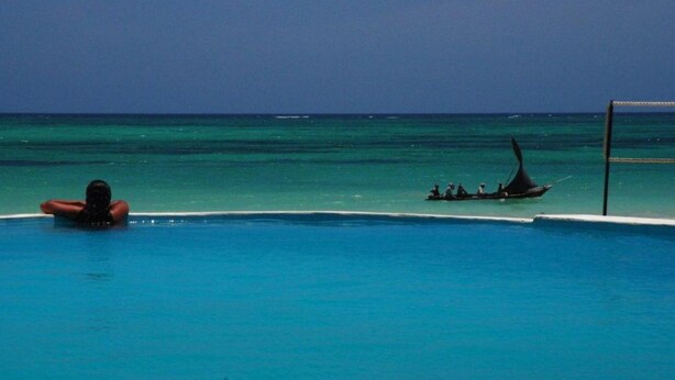 A pool with clear blue water, a person relaxing in the water, and a boat with people in the background. - Kusini Beach Cottages (Diani Beach)