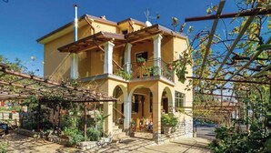 A two-story building with a balcony, arched windows, and a garden area with plants and a pergola. - Apartment Kumicic (Opatija)