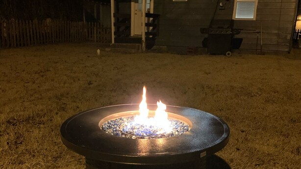 Ein rundes Feuerbecken im Freien mit Flammen, umgeben von einer Rasenfläche, mit einem Haus im Hintergrund.
