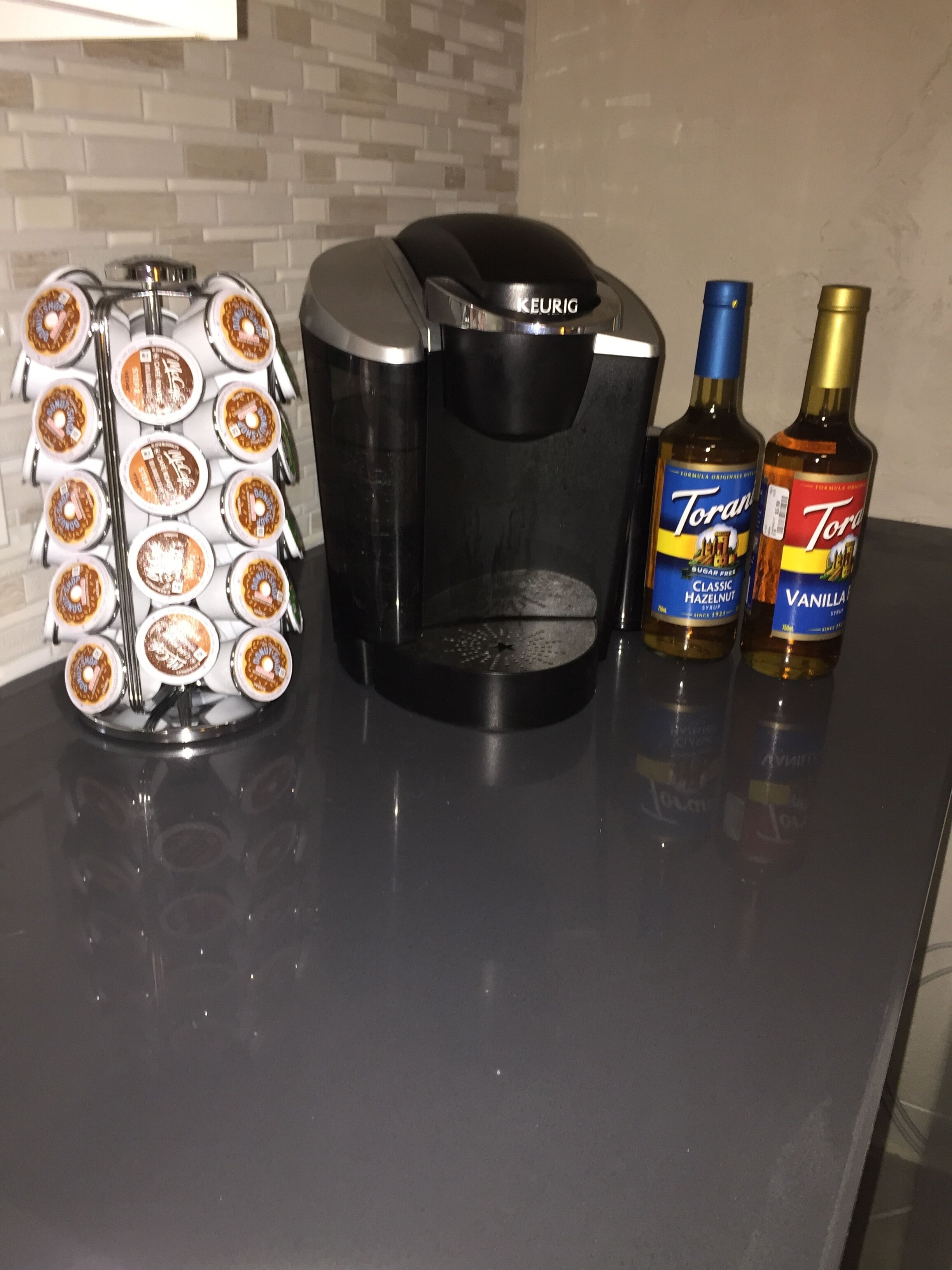 Une machine à café, deux bouteilles de sirop Torani et un distributeur de dosettes de café, le tout posé sur un plan de travail.