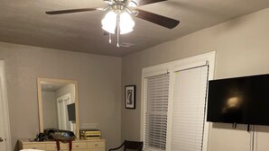 Ein Schlafzimmer mit einem Bett, einem Deckenventilator, einem Schminktisch, einem Sessel, einem Fernseher und einem Fenster mit Jalousien.