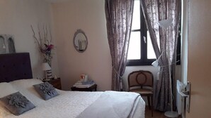 A bedroom with a bed, a nightstand, a chair, a lamp, and a window with curtains. - CHAMBRE D'HÔTE CONFORTABLE (Voisins-le-Bretonneux)