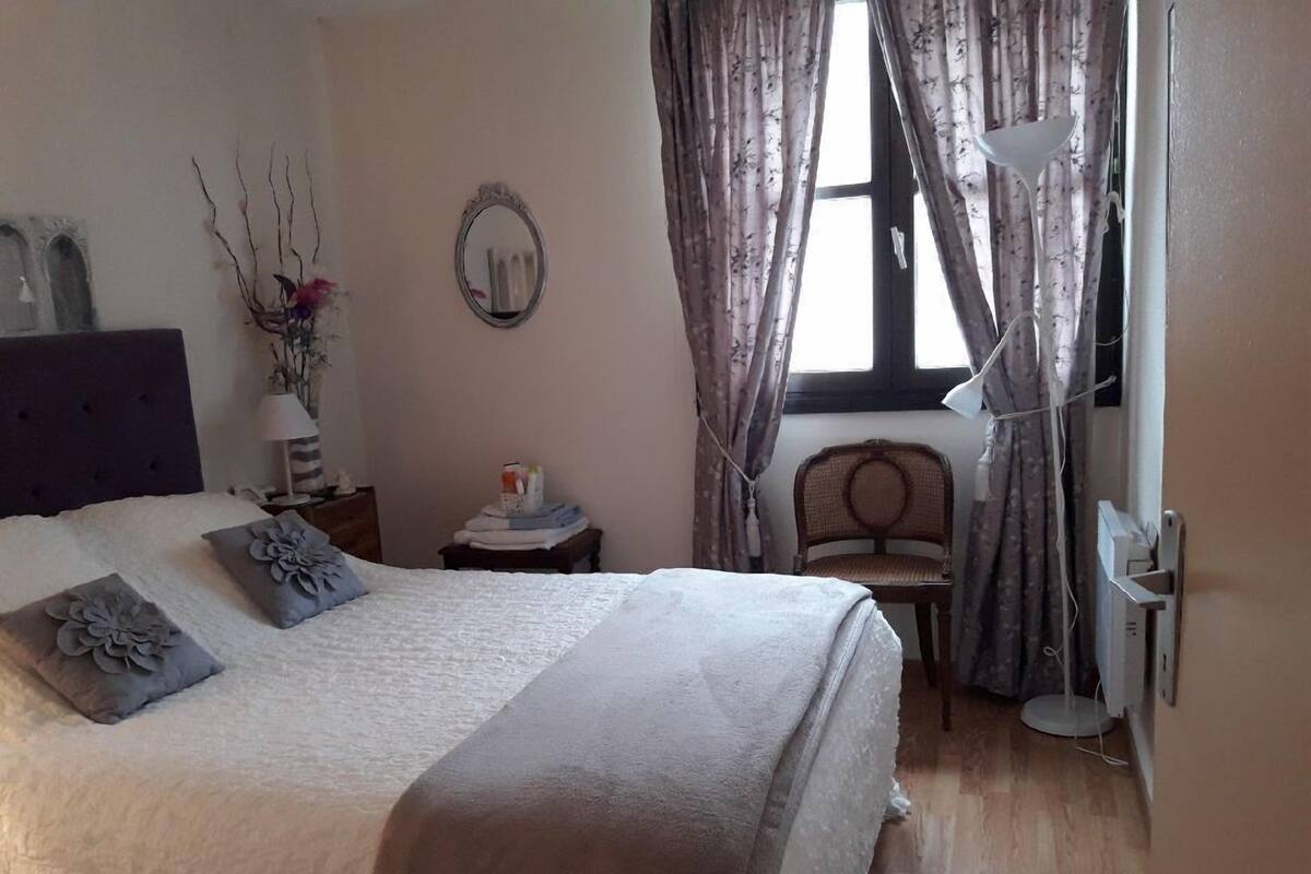 Ein Schlafzimmer mit einem Bett, einem Nachttisch, einem Sessel, einer Lampe und einem Fenster mit Vorhängen.