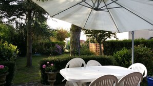 An outdoor patio with a white table and chairs under a large umbrella, surrounded by lush greenery and a brick wall. - CHAMBRE D'HÔTE CONFORTABLE (Voisins-le-Bretonneux)