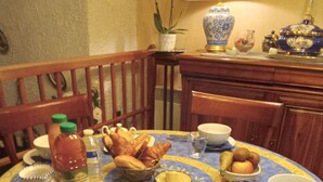 A dining table with a floral tablecloth, surrounded by chairs. There are various food items and condiments on the table, including bread, jars of preserves, and a bottle of water. - CHAMBRE D'HÔTE CONFORTABLE (Voisins-le-Bretonneux)