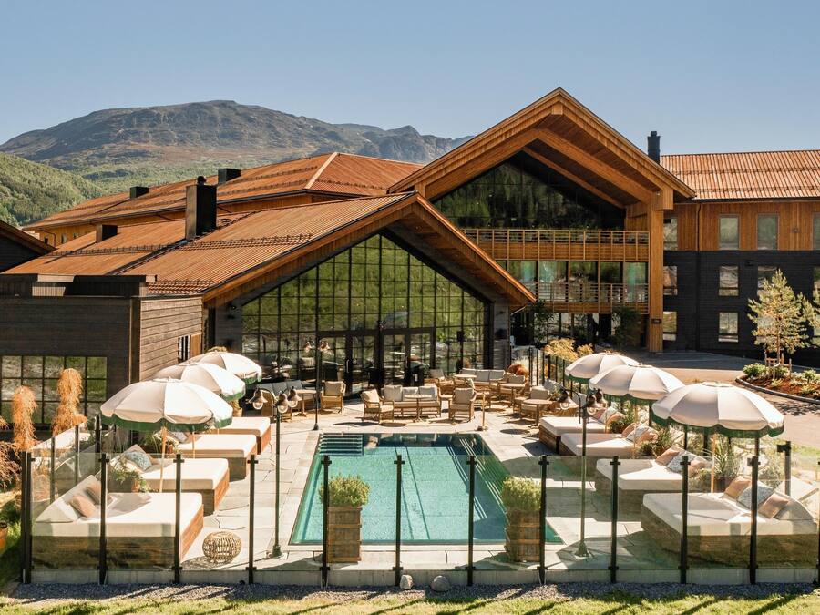 Un hotel moderno con un amplio área de piscina, mobiliario exterior y vistas a la montaña.